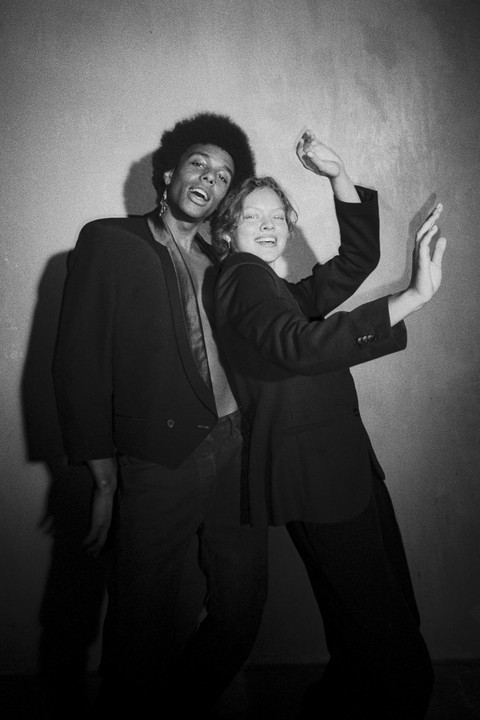 A man and a woman in blazers pose playfully against a wall.