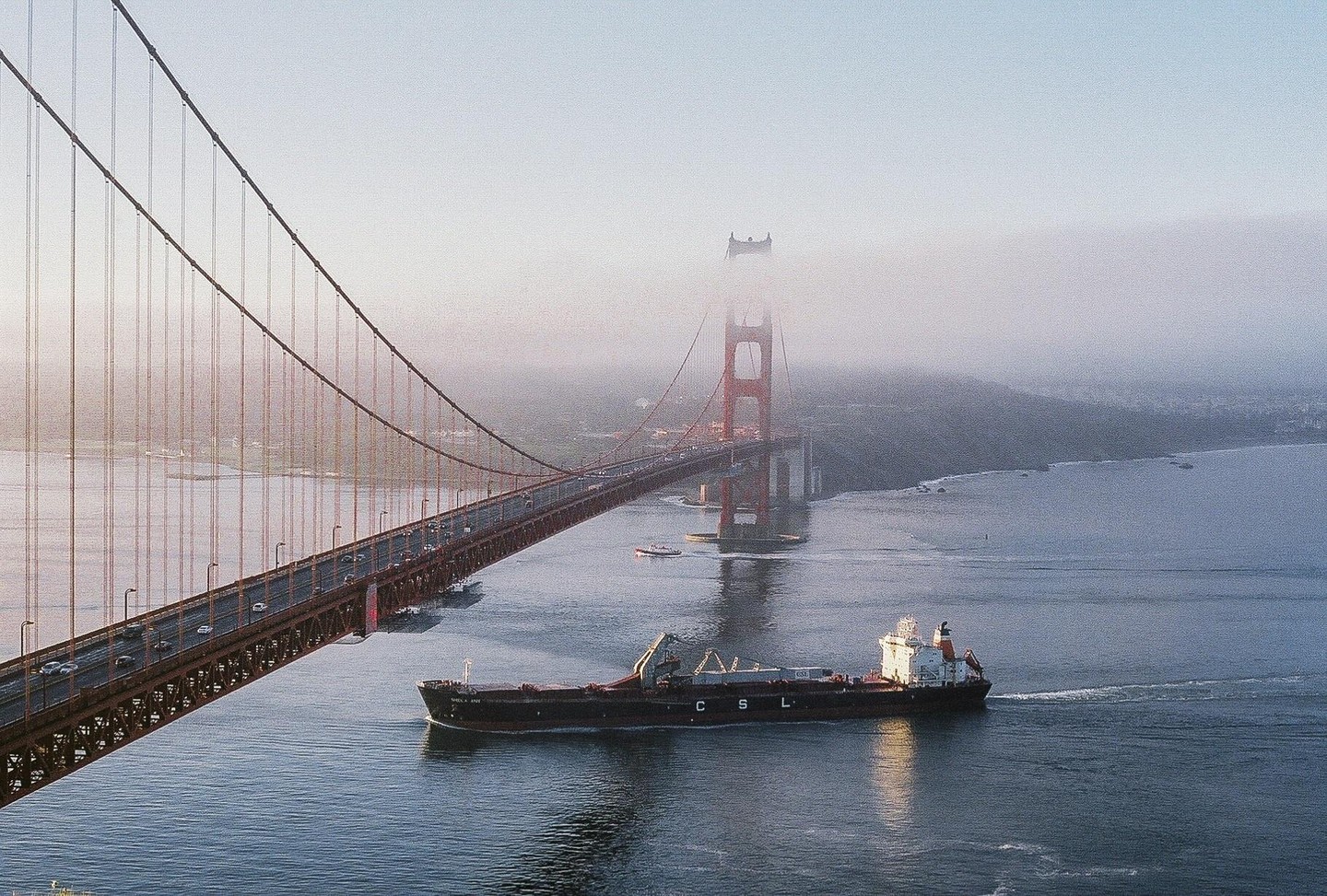 A cargo ship passes under the Golden Gate Bridge, which is partially obscured by fog.