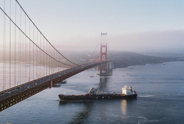 A cargo ship passes under the Golden Gate Bridge, which is partially obscured by fog.