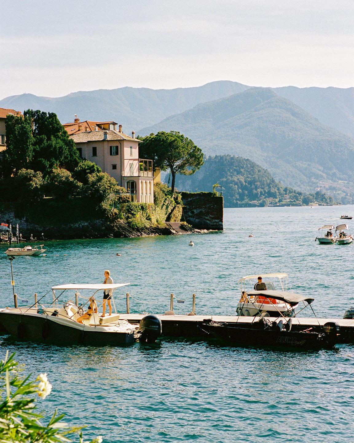 A woman stands on a boat docked at a pier on a lake with mountains and buildings in the background.