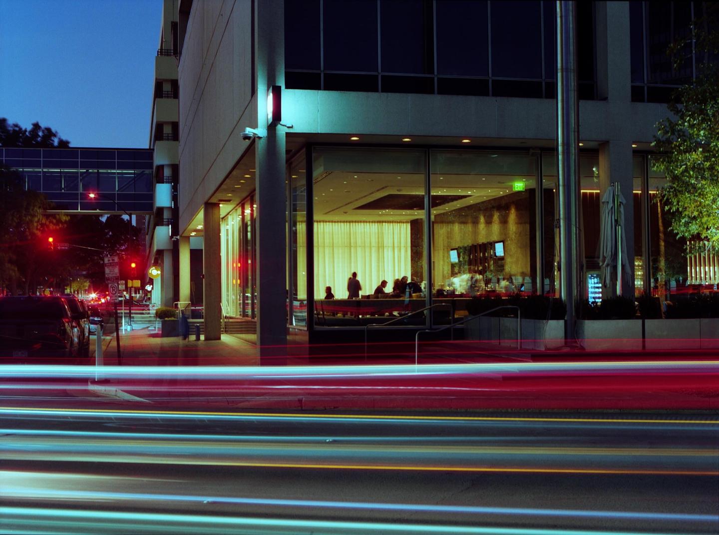 A street at night shows light trails from vehicles and a building with people visible through its large windows.