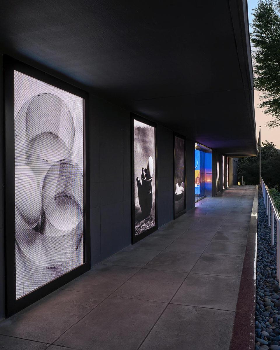 A series of illuminated artworks are displayed along a dark building facade with a tiled walkway and a bed of river rocks.