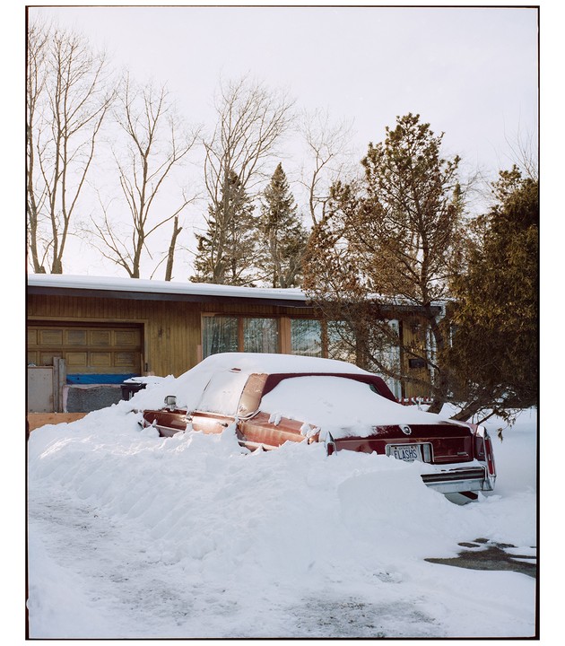 A red car is buried in deep snow in front of a house with a garage and bare trees in the background.