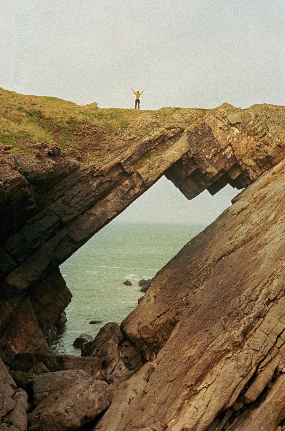 A person stands atop a natural rock arch overlooking the ocean.