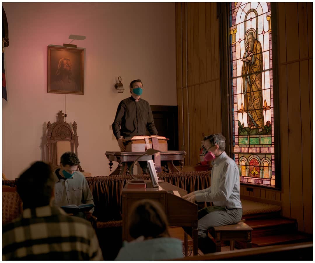 A man in a face mask stands at a pulpit in a church while another man plays the organ and congregants sit in the pews.
