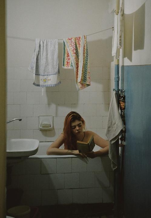 A woman with red hair reads a book while sitting in a bathtub in a tiled bathroom with towels hanging on a line.