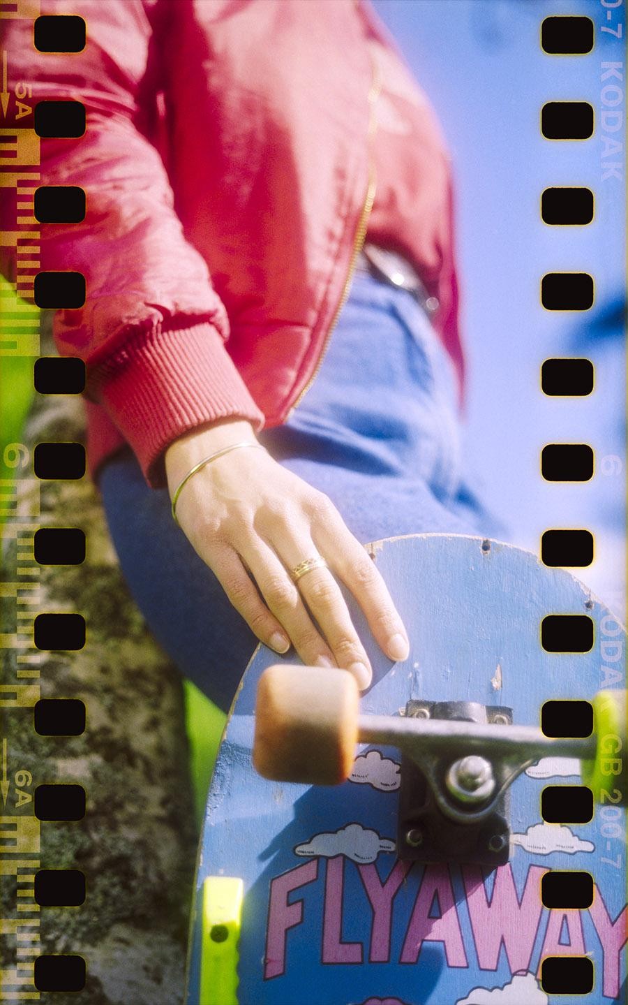 A person in a red jacket and blue jeans holds a blue skateboard with a "FLYAWAY" logo.