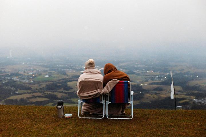 A couple sits in chairs on a grassy hill, looking out at a hazy landscape with distant lights.
