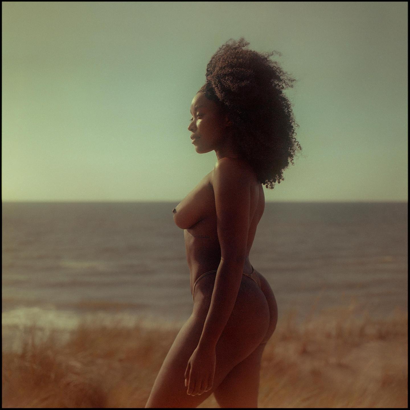 A woman with curly hair stands in profile on a grassy dune overlooking the ocean.