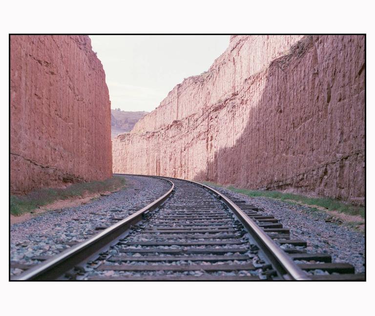 A set of train tracks curves through a narrow canyon with tall, reddish rock walls.