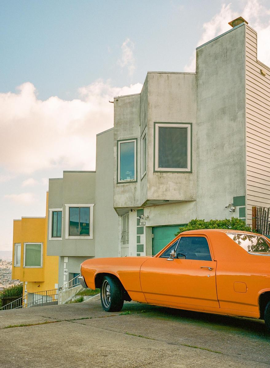 An orange classic car is parked on a sloped street in front of several houses with a view of a city in the background.