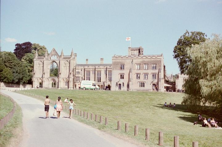 A group of people walk along a paved path towards a large stone building with an arched facade and a flag flying from a tower.