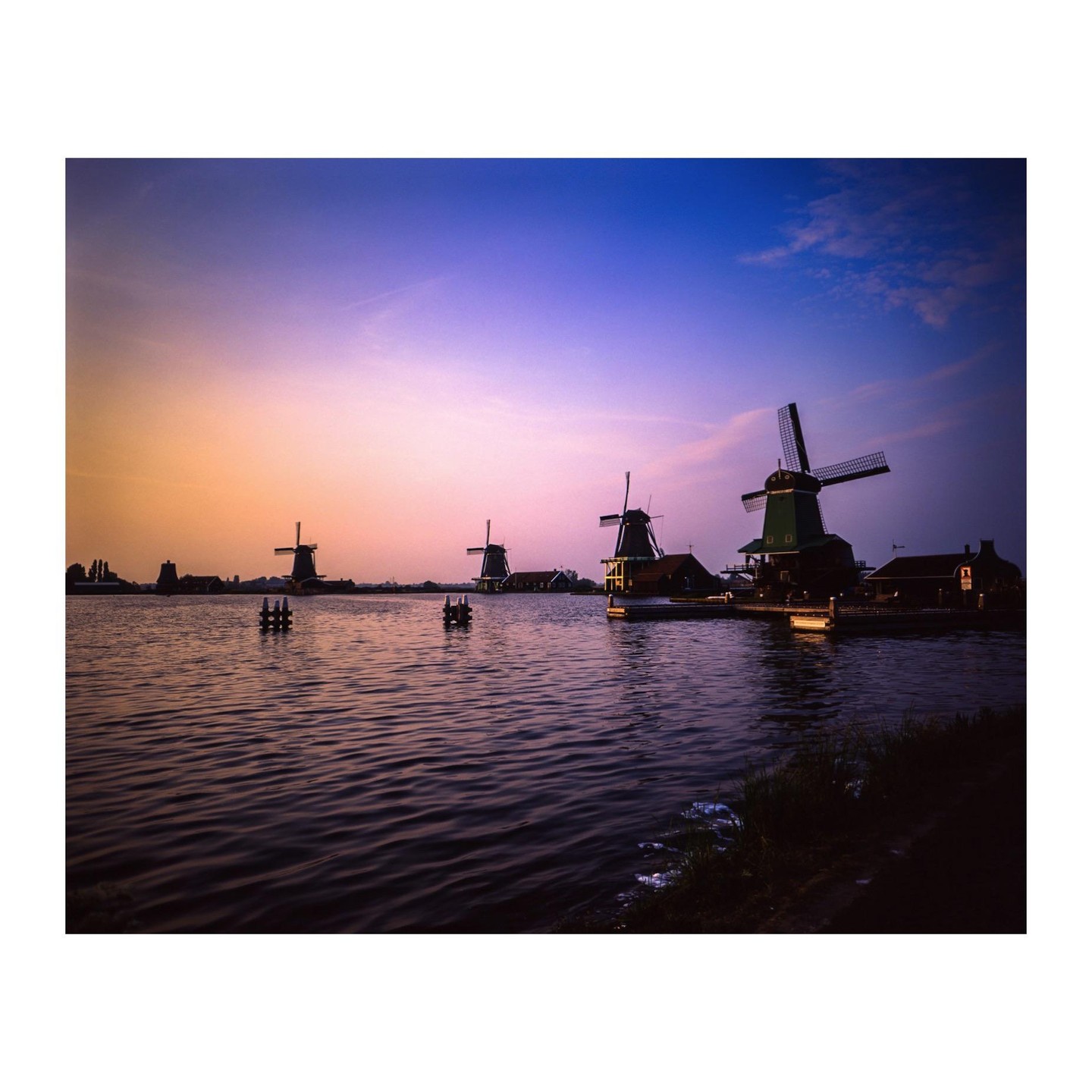 Several windmills stand along a body of water under a colorful sky at dusk.