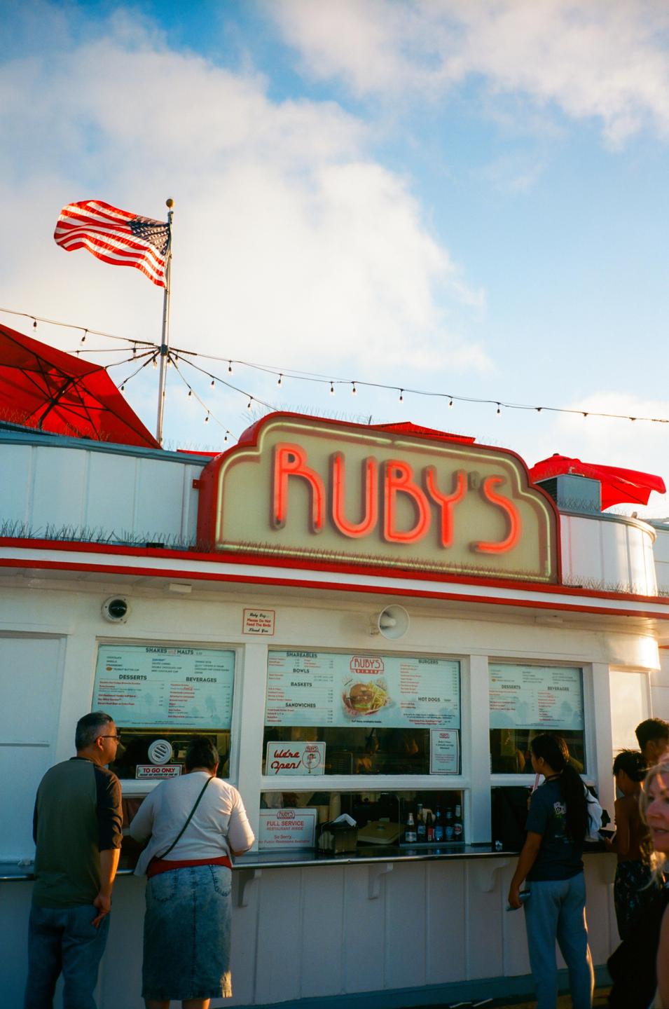 A group of people stand in line at a Ruby's restaurant with an American flag flying above.