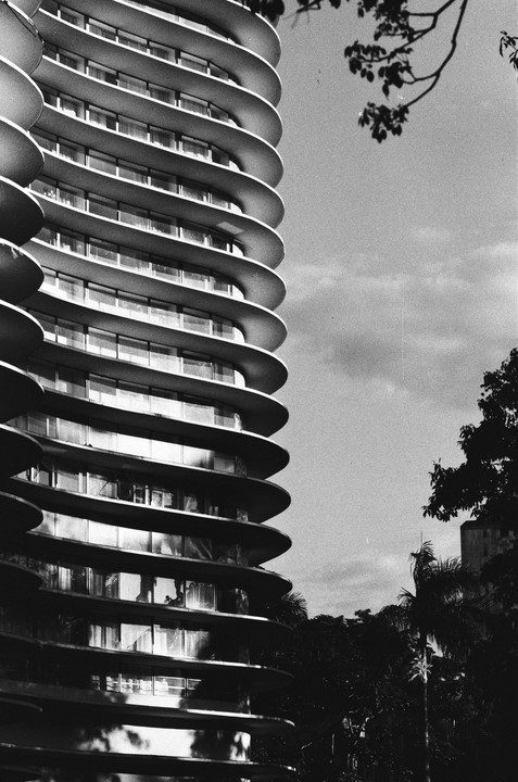 A tall, curved building with many balconies stands next to trees under a cloudy sky.