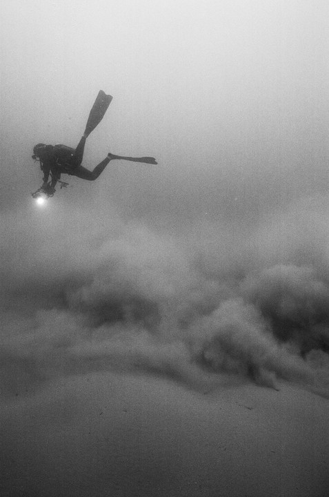 A diver with a light attached to their camera floats above a cloud of sand underwater.