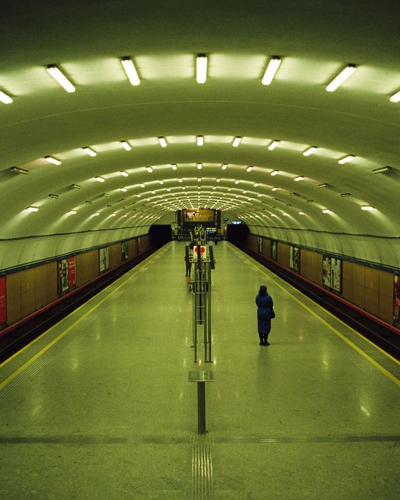 A person stands on a subway platform with a long, arched ceiling illuminated by many fluorescent lights.