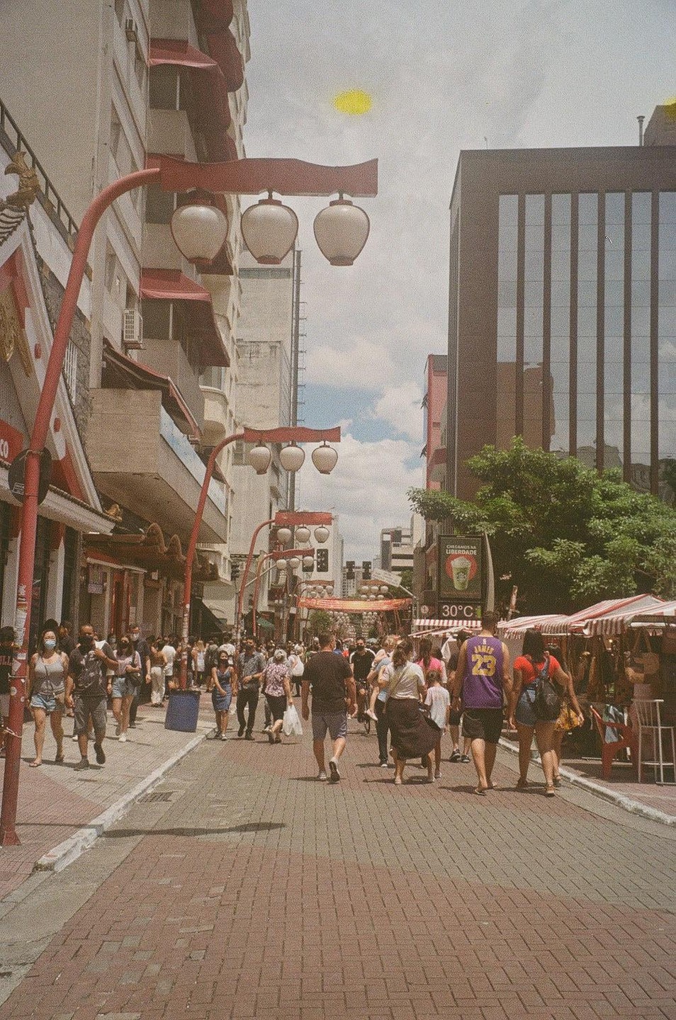 A street scene shows many people walking on a brick road lined with buildings and red decorative streetlights.