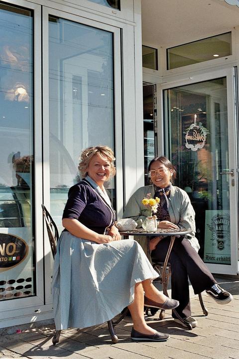Two women sit at a small table outside a cafe with glass windows, smiling at the viewer.