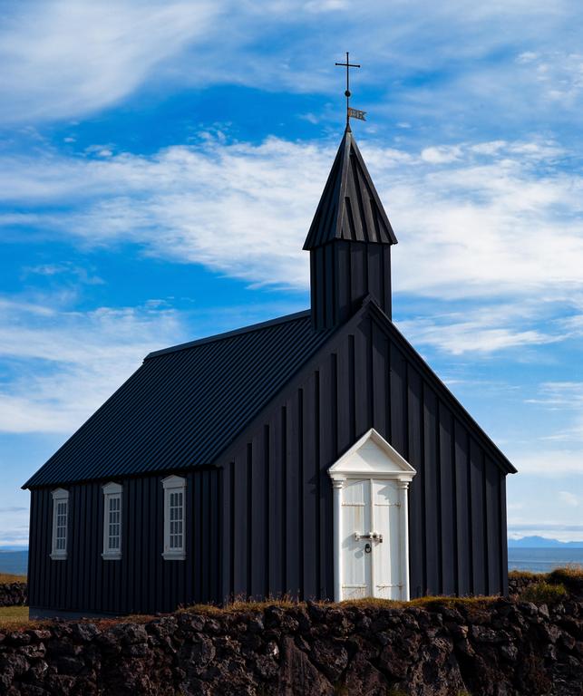 A black church with a white door and steeple stands on a stone wall under a blue sky with white clouds.