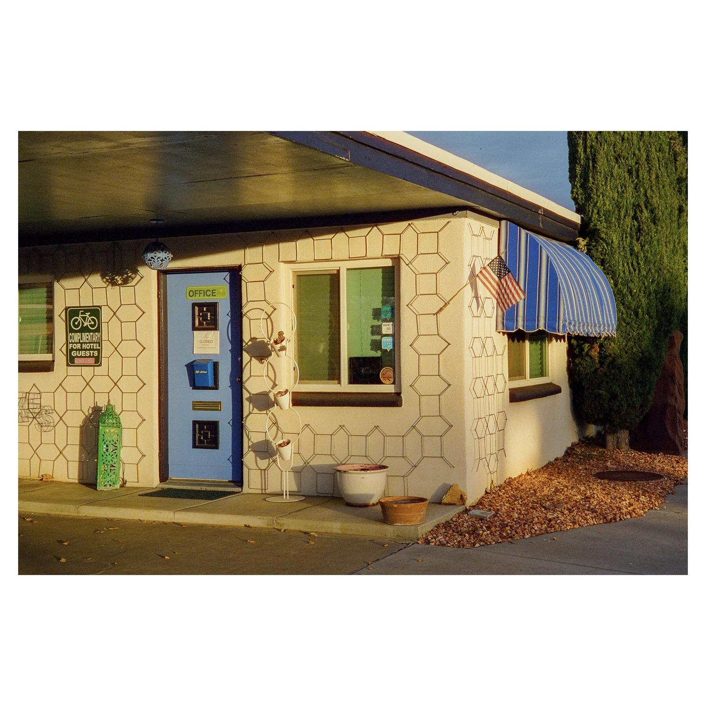The office of a motel with a blue door and a striped awning is illuminated by the morning sun.