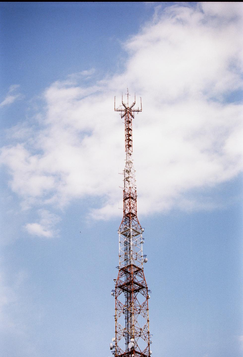 A tall red and white transmission tower stands against a blue sky with white clouds.
