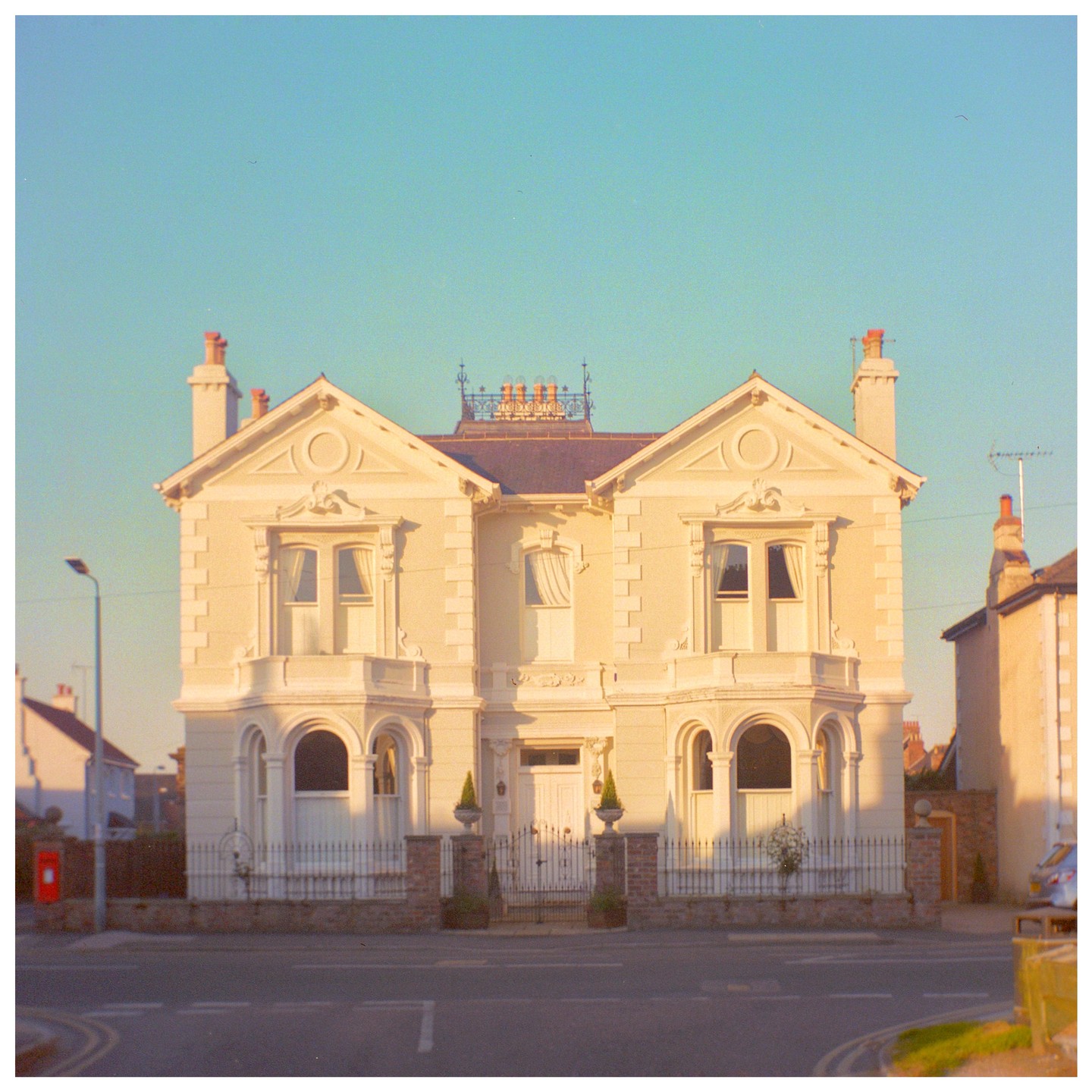 A large, ornate house with a symmetrical facade stands at a street corner under a clear sky.