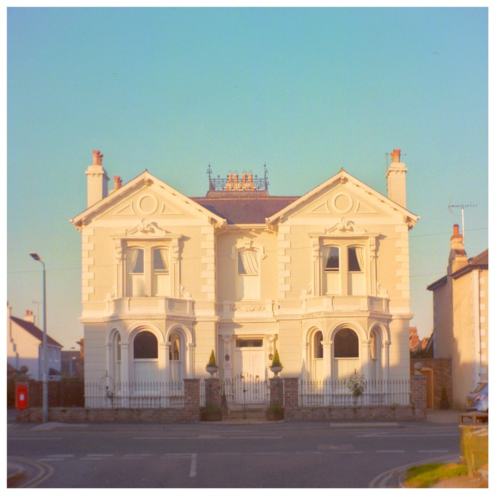A large, ornate house with a symmetrical facade stands at a street corner under a clear sky.