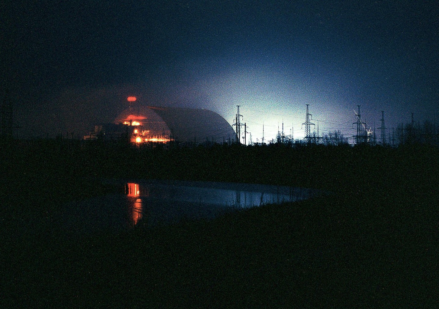A nuclear power plant with a large containment dome glows orange and blue at night, with power lines silhouetted against the sky and reflections in a body of water.