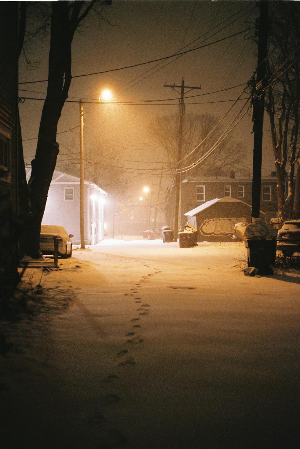 Footprints lead down a snow-covered alley illuminated by streetlights at night.