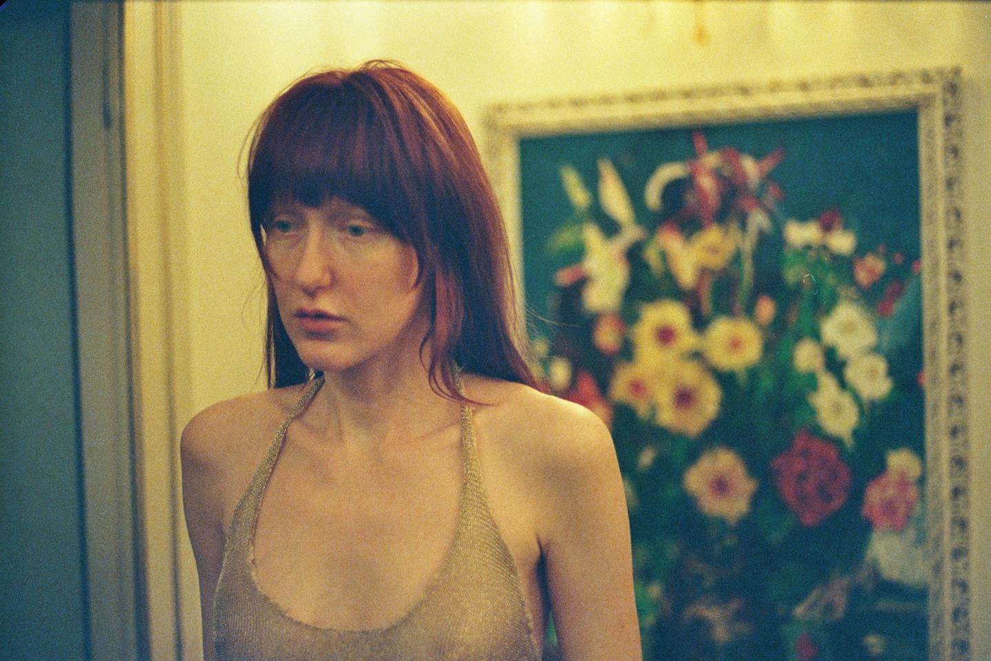 A woman with red hair and a gold top looks to the left of the frame, with a painting of flowers in the background.