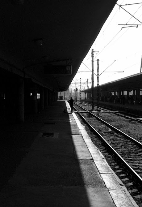 A person stands on a train platform next to railroad tracks, with a strong shadow cast across the platform.