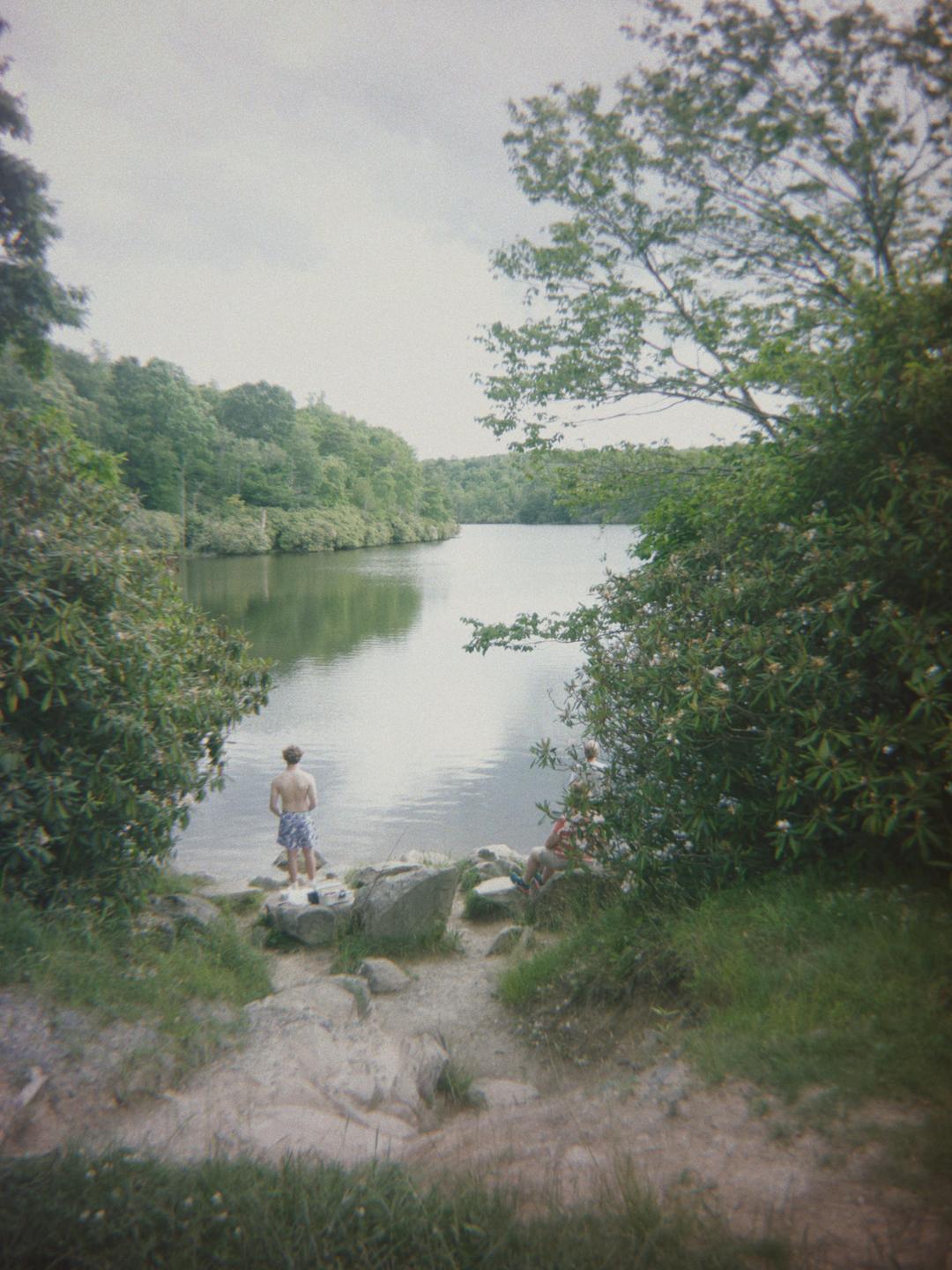 A man stands at the edge of a lake surrounded by lush green trees and bushes, with another person partially visible behind a bush.