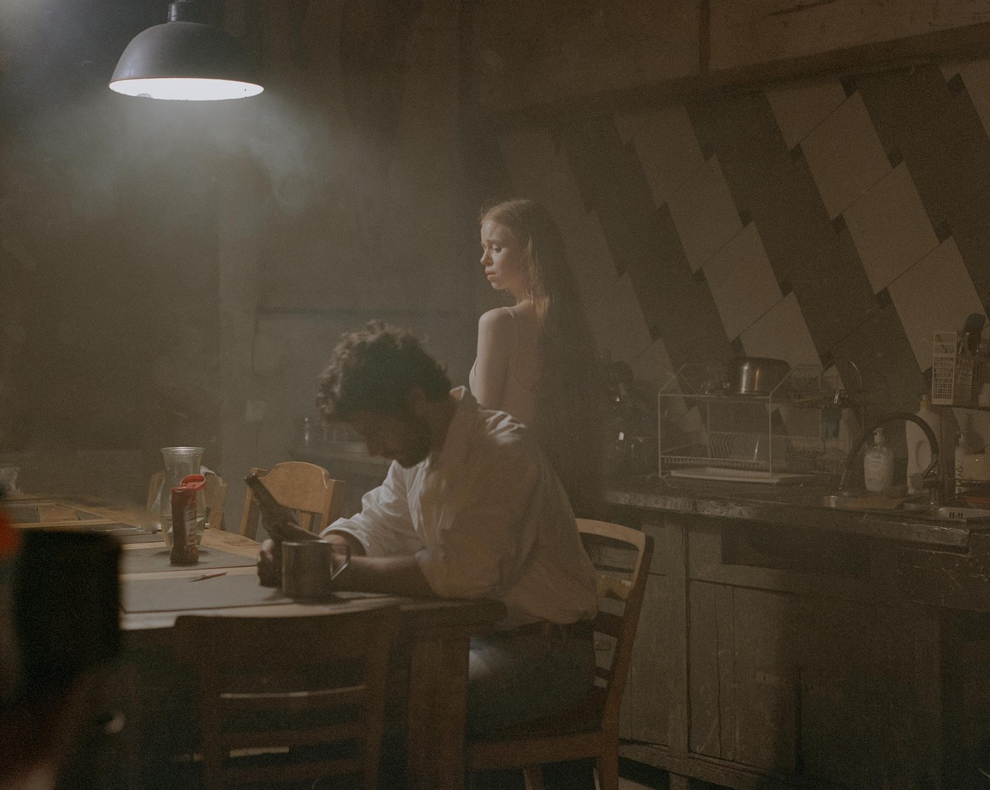 A man sits at a table while a woman stands behind him in a dimly lit room.