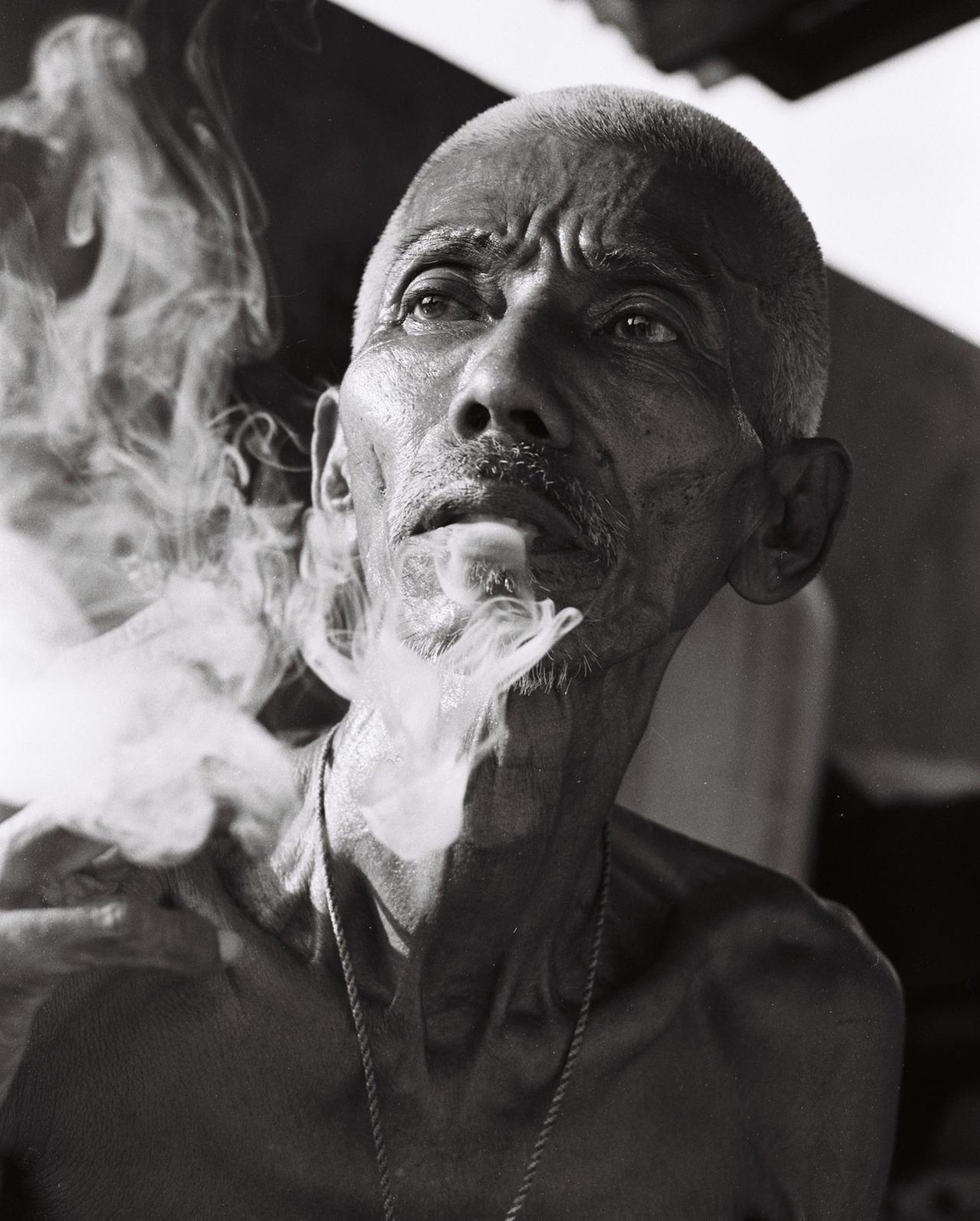 A man with a shaved head and a thin mustache exhales a cloud of smoke while looking up.