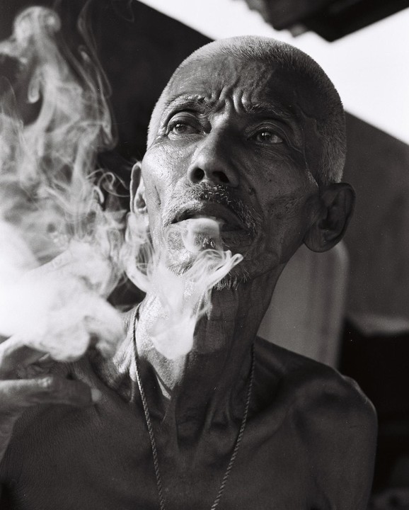 A man with a shaved head and a thin mustache exhales a cloud of smoke while looking up.