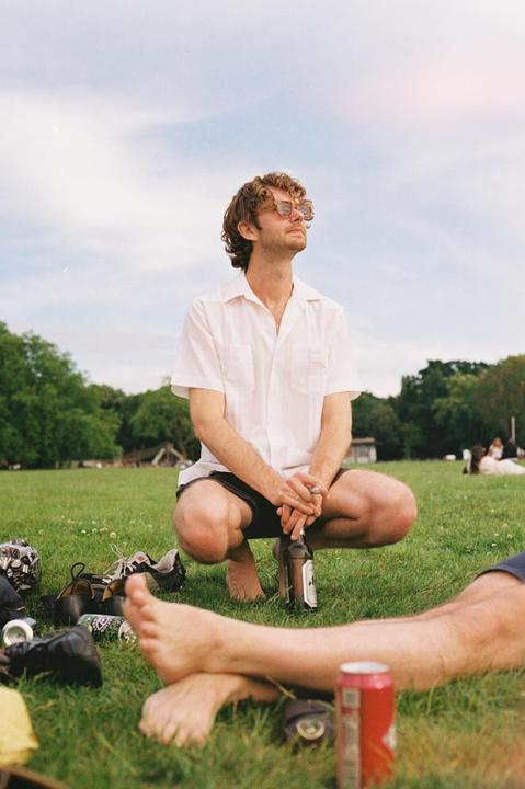 A man wearing sunglasses and a white shirt crouches in a grassy field, holding a bottle.