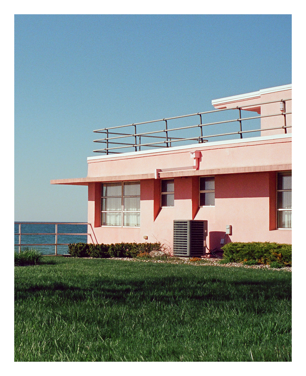 A pink building with a flat roof and railing stands next to a body of water, with green grass in the foreground.