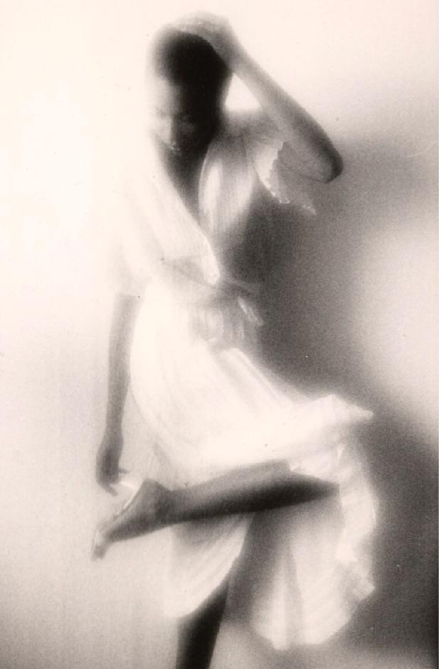 A woman in a light-colored dress poses with one leg raised and her hand on her head.