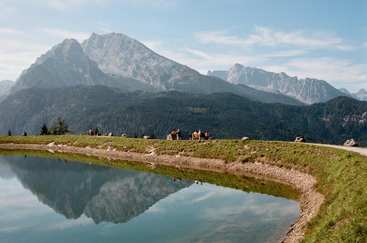 A mountain range with a forest at its base is reflected in a lake, with people sitting on a grassy bank in the middle ground.