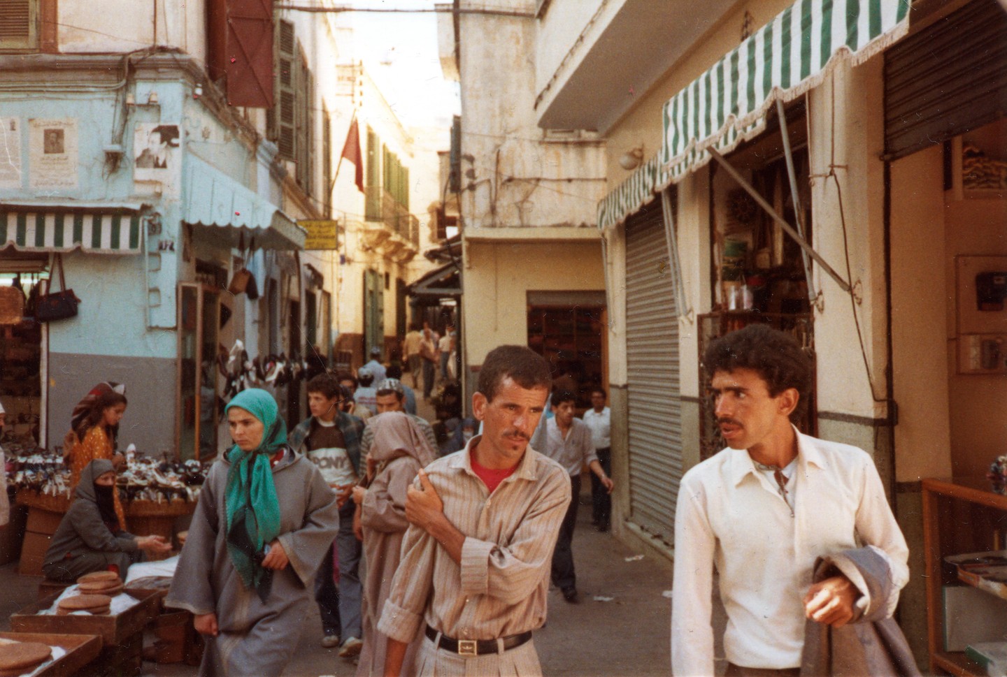 A busy street in Tangier is filled with people, some walking and some tending to market stalls.