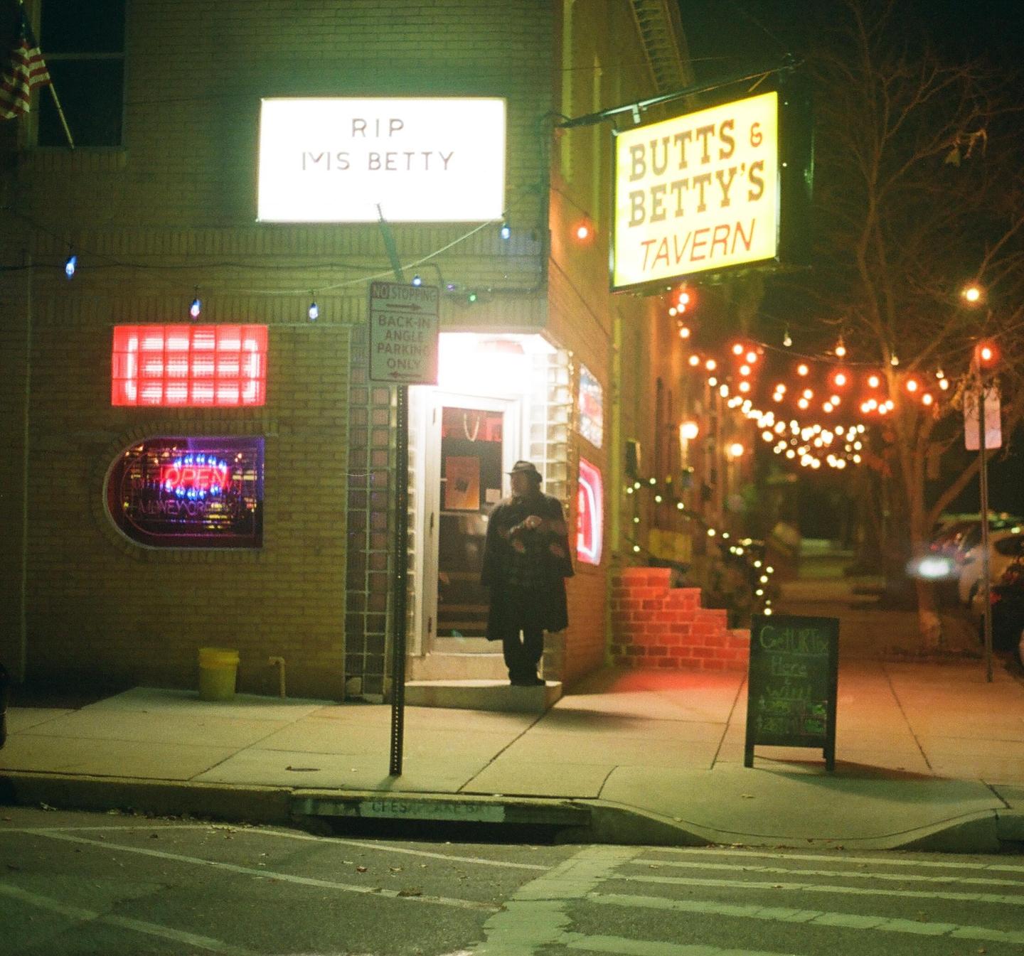 A person stands outside a tavern with glowing signs and string lights at night.
