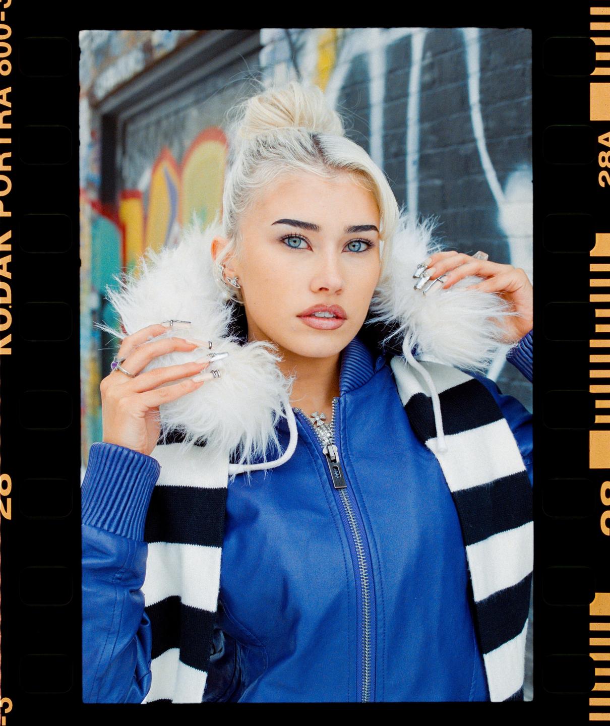 A woman with blonde hair and blue eyes poses for a portrait while wearing a blue jacket and a striped scarf.