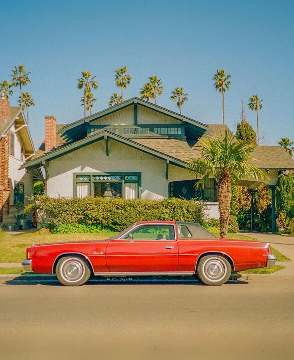 A red car is parked on a street in front of a house with palm trees in the background.