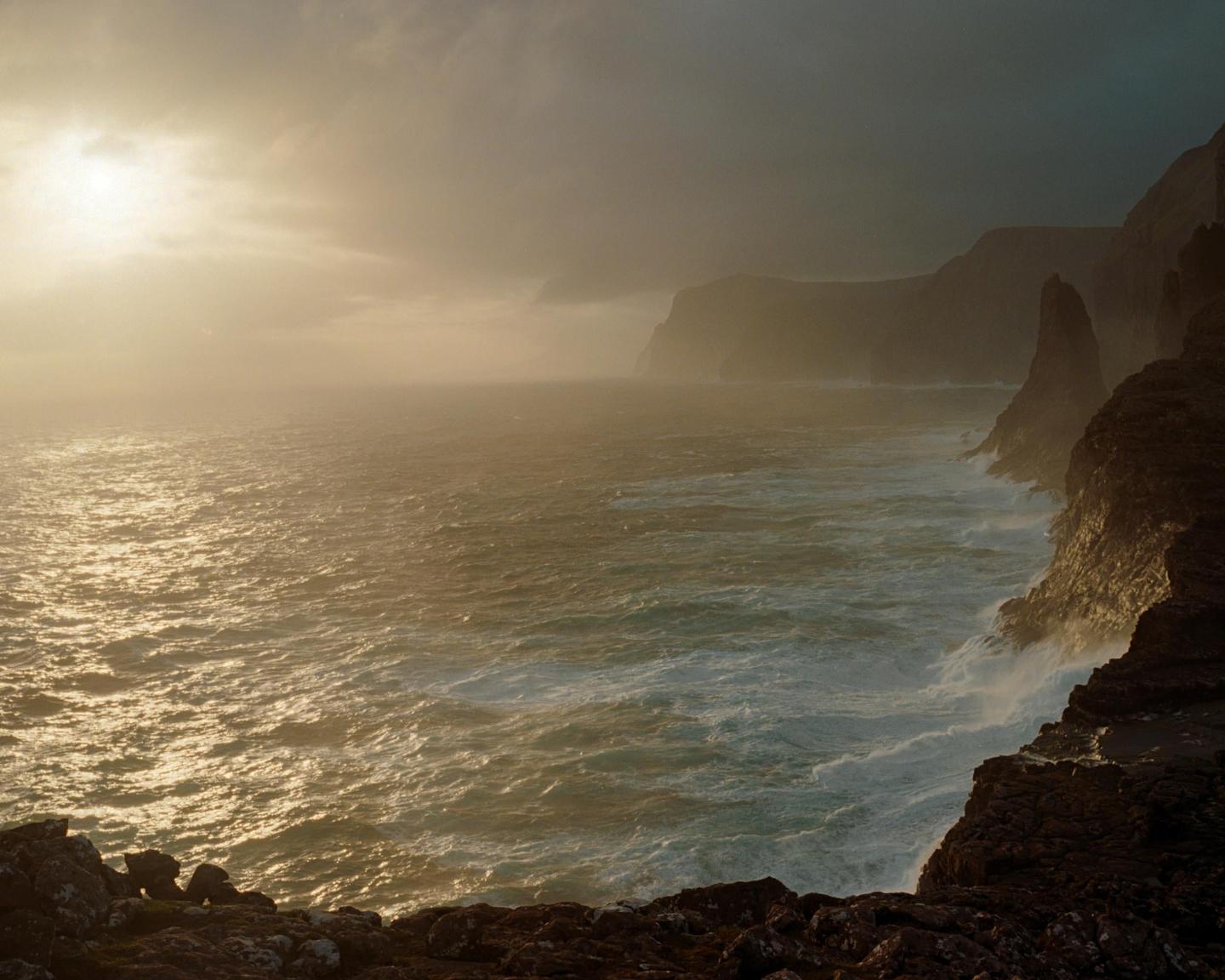 Waves crash against a rocky coastline under a hazy sky with a bright sun.