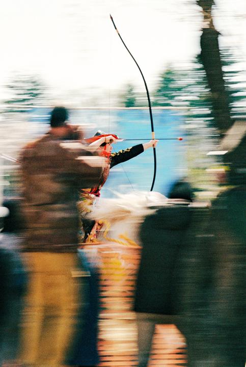 A person in traditional attire aims a bow and arrow while riding a horse, surrounded by blurred spectators.