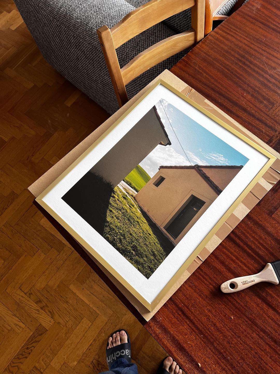 A framed print of a house and a grassy field sits on a wooden table next to a pair of feet in sandals.