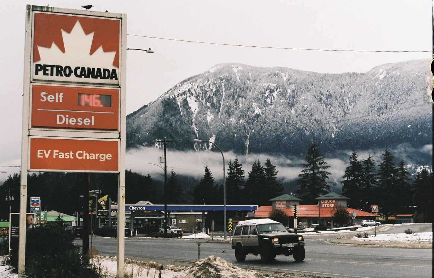 A Petro-Canada sign stands beside a road with a snow-covered mountain and a gas station in the background.