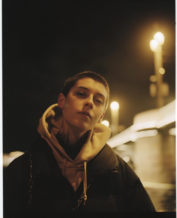 A woman with short hair and a hoodie looks directly at the viewer at night, with streetlights in the background.