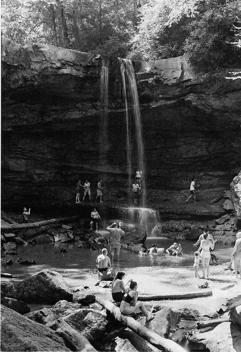 A group of people are gathered around a waterfall and swimming in the pool below it.